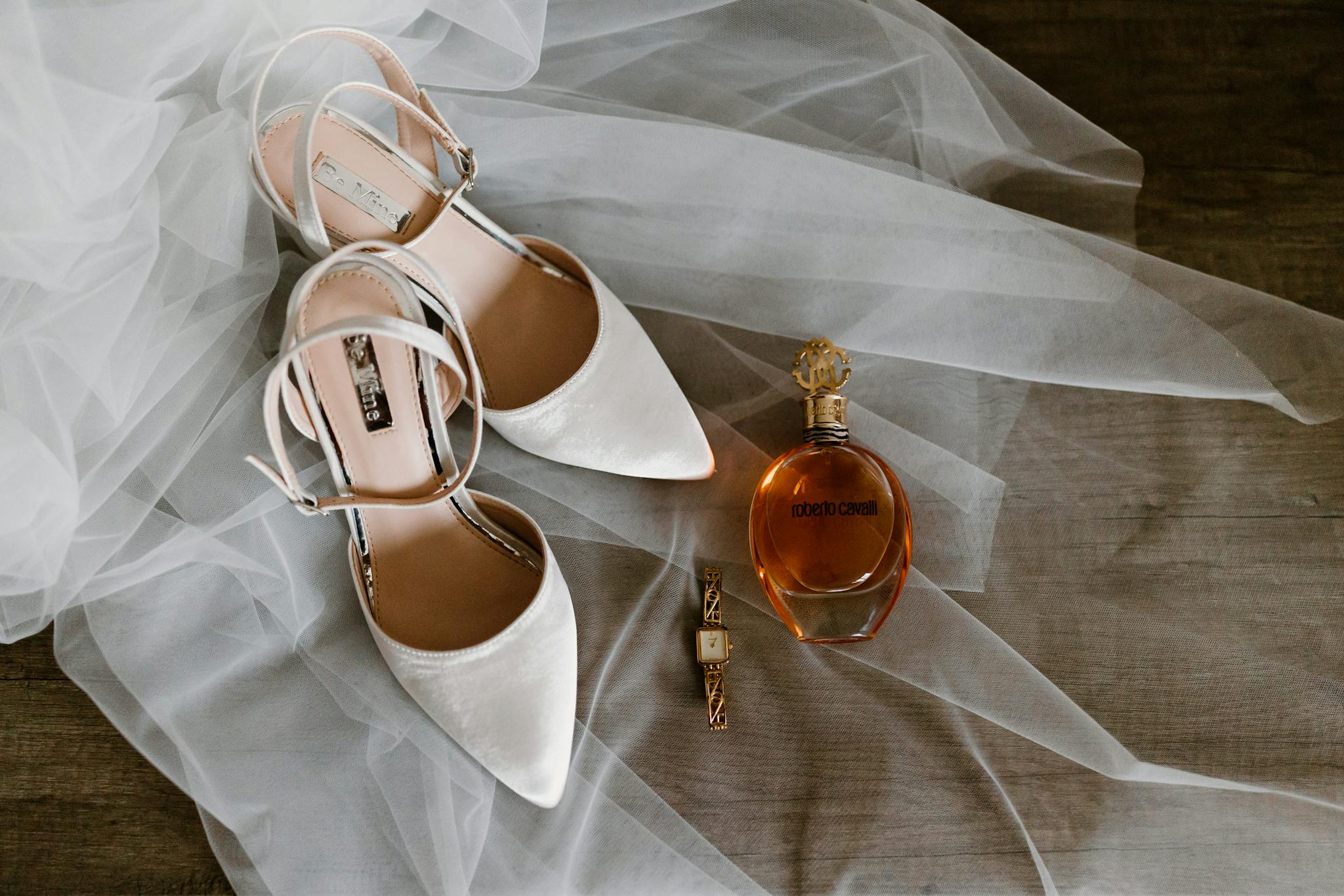 Chic wedding essentials displayed on sheer fabric include white pointed shoes, a stylish watch, and designer perfume.