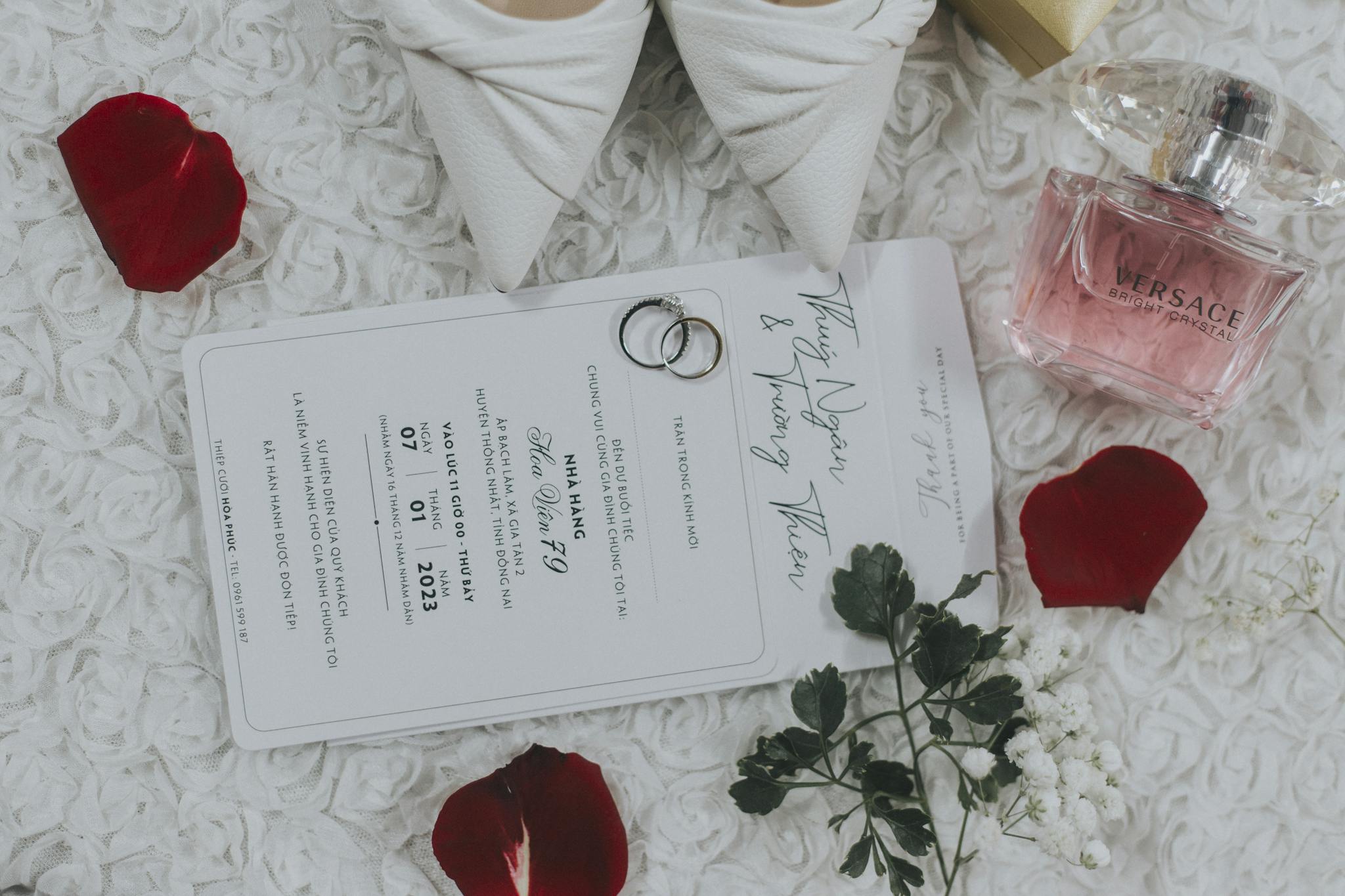 Flat lay of a wedding invitation with rings and perfume, surrounded by petals.