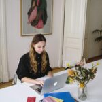 Young woman using a laptop at a desk with documents and flowers, in a bright, modern workspace.