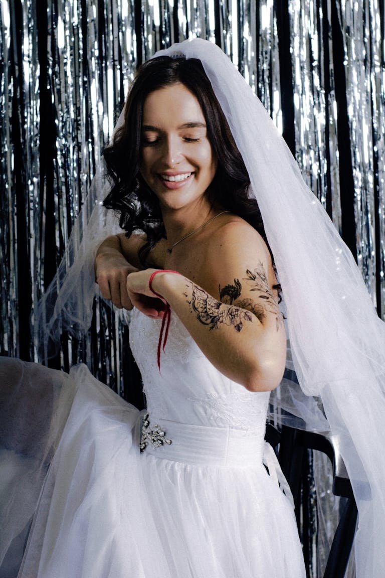 A joyful bride in a white wedding gown with tattoos and a veil, smiling indoors.