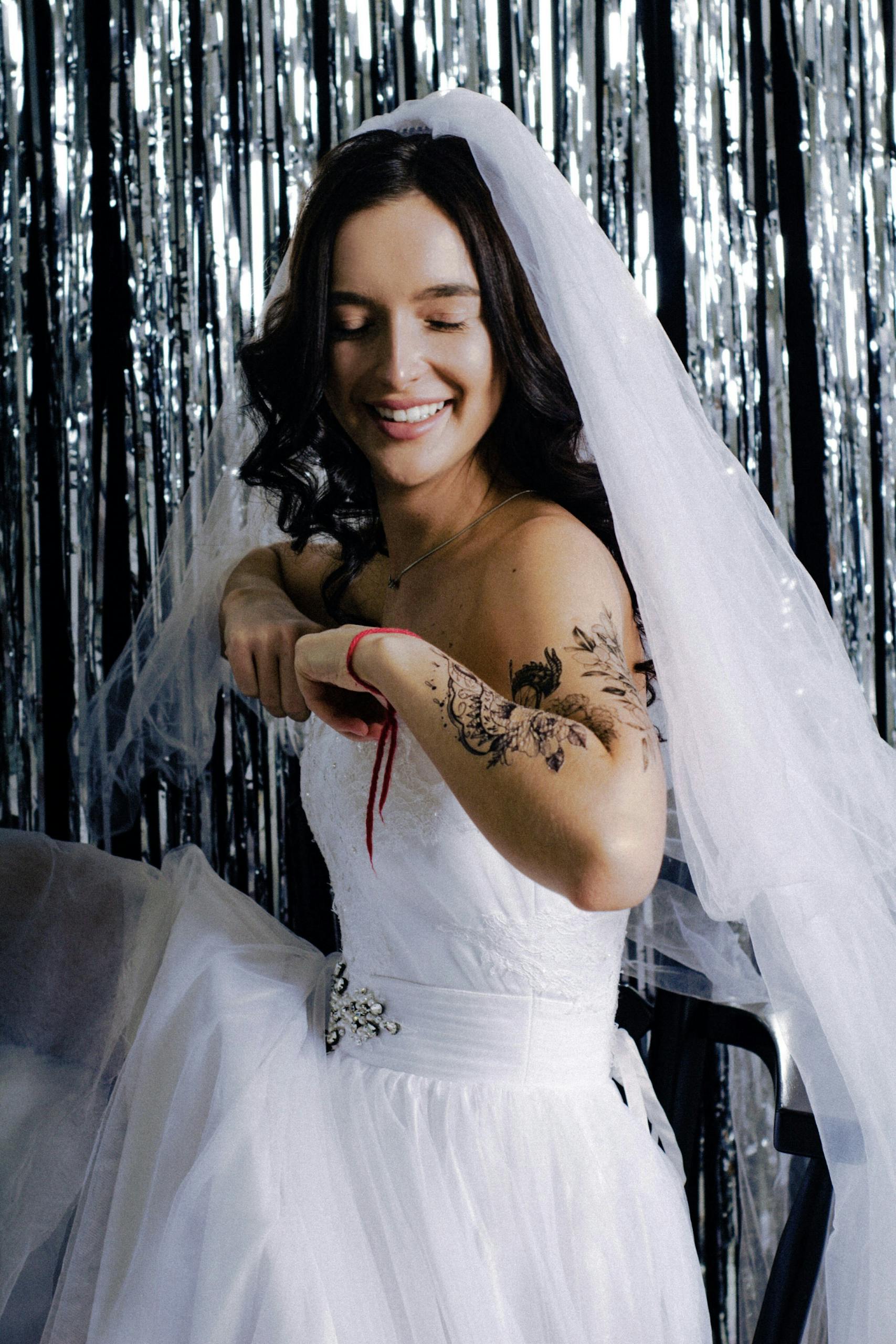A joyful bride in a white wedding gown with tattoos and a veil, smiling indoors.